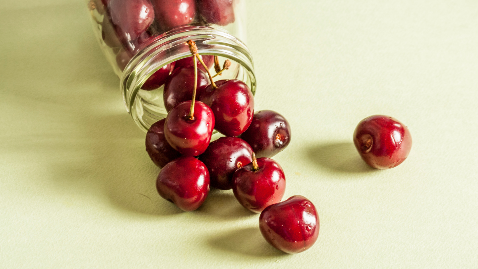 red cherries falling out of glass jar on cream background